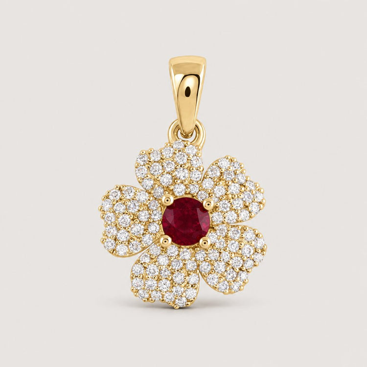 The 1/4CTW Diamond Flower Pendant in 14K Yellow Gold showcases five diamond-studded petals and a central red gemstone, creating an elegant floral design against a plain warm off-white studio background.