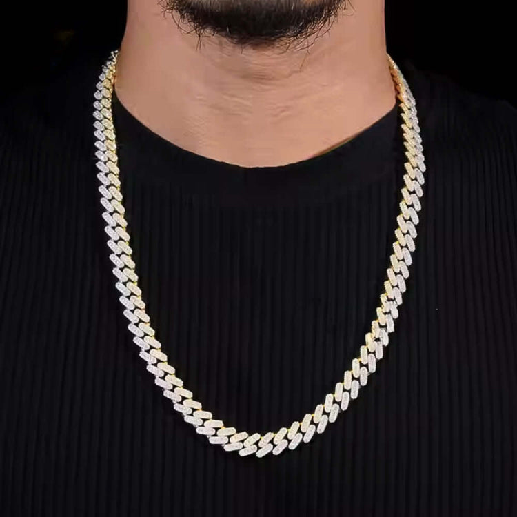 A man wears the Moissanite Cuban Link Chain - 10mm, a thick gold and silver necklace, over a ribbed black shirt, shown from shoulders to chin against a dark background.