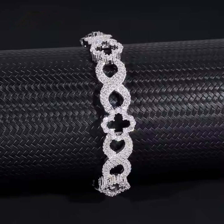 The Infinity Clover Moissanite Bracelet - 11mm features heart-shaped links encrusted with sparkling stones, elegantly displayed on a textured black cylindrical surface.