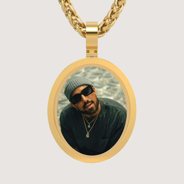 The 14K Gold Classic Oval Picture Pendant features a photo of a man in a gray beanie and sunglasses with layered necklaces, looking upward. This striking pendant is a unique statement piece.