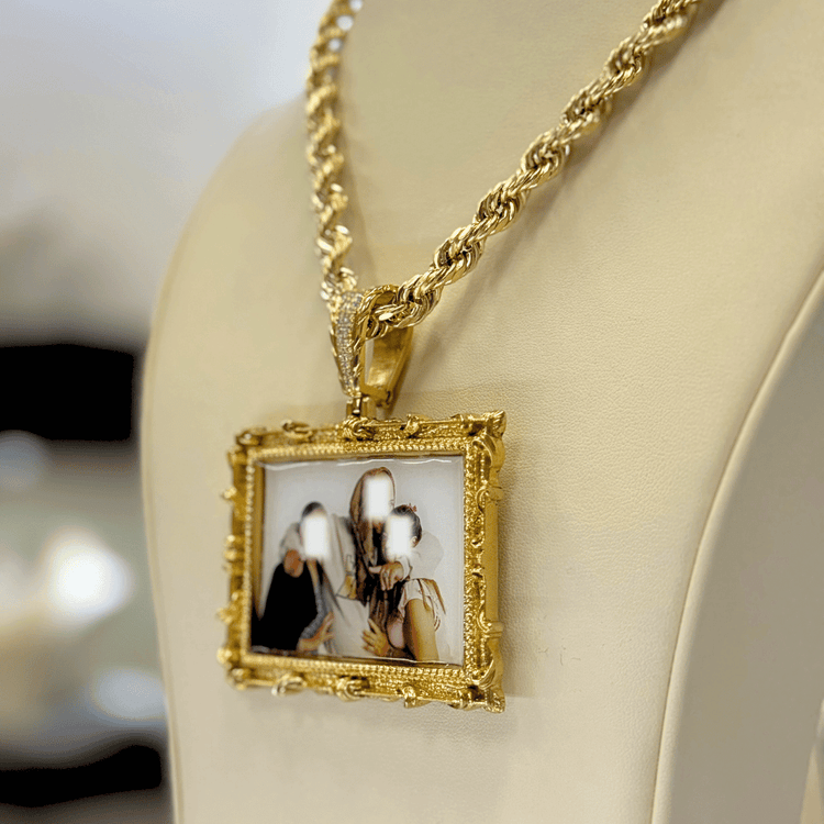 The 14K Gold Diamond Bail Big Frame Picture Pendant features an ornate rectangular gold setting with a blurred photo of three people, hanging from a gold rope chain and elegantly displayed on a cream-colored stand.