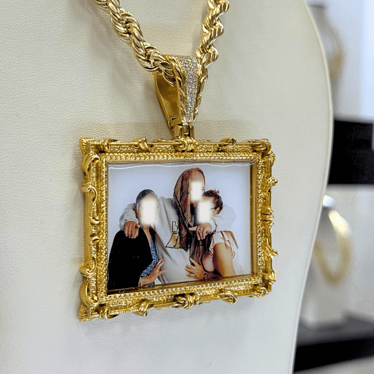The 14K Gold Diamond Bail Big Frame Picture Pendant features a large, ornate rectangular design displaying a blurred family photo of three people on a white jewelry stand.