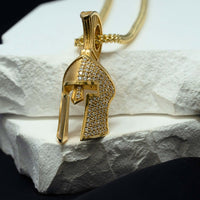 The Half-Face Diamond Spartan Helmet Pendant in 14K gold, featuring sparkling stones and a helmet design, hangs from a gold chain and rests on textured white stone slabs against a dark background.