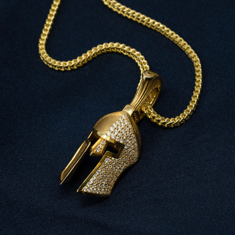The Half-Face Diamond Spartan Helmet Pendant in 14K Gold features a helmet-shaped design with sparkling stones, showcased on a dark fabric background.
