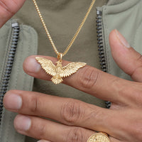 Someone holds up the 14k Yellow Gold Eagle Pendant, featuring a detailed bird design, while wearing a gold ring and a light green jacket with a visible zipper.