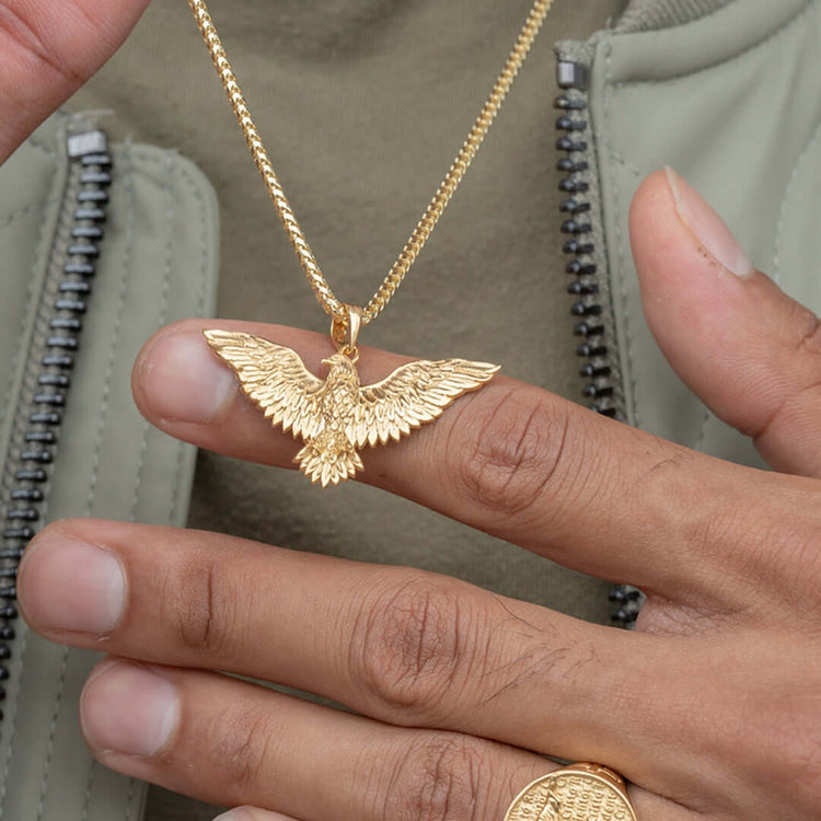 Someone holds up the 14k Yellow Gold Eagle Pendant, featuring a detailed bird design, while wearing a gold ring and a light green jacket with a visible zipper.