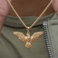 A close-up of the 14k Yellow Gold Eagle Pendant, held by two fingers. The background features a green shirt and silver zipper, emphasizing the fine craftsmanship and bold style of this striking eagle jewelry piece.