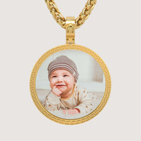 A 14K Gold Greek Key Round Picture Pendant featuring a photo of a smiling baby in a striped hat and patterned outfit, lying on their stomach against a light background.