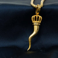 The Italian Horn Pendant – 14K Gold, shaped like a cornicello with an ornate crown top, hangs from a gold chain against a dark textured fabric background.