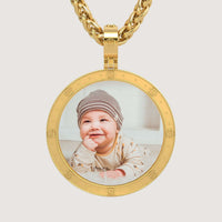 The 14K Gold Round Engraved Border Picture Pendant displays a photo of a smiling baby in a striped hat and light outfit, lying down with a hand under their chin—a charming, personalized accessory.