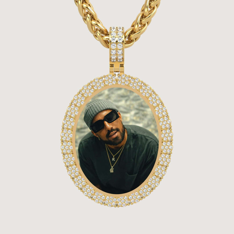 The 14K Gold Scalloped Oval Diamond Photo Pendant features a diamond-encrusted oval frame and showcases a photo of a man in a beanie, sunglasses, and layered necklaces gazing up at the camera.