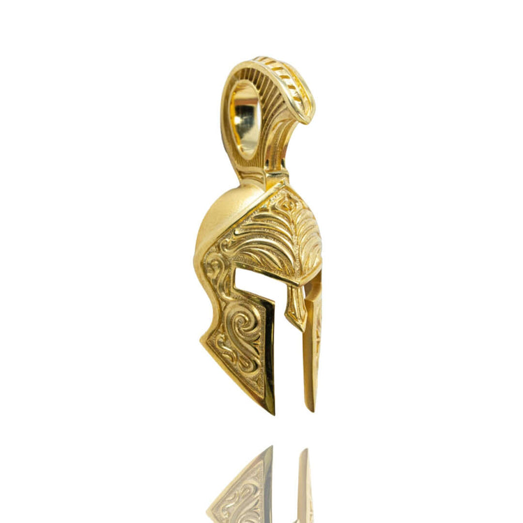 The Spartan Helmet Pendant - 14k Solid Gold is shaped like an ancient Greek warrior’s helmet, features detailed engraved patterns, and includes a loop for a chain. It’s photographed against a white background.