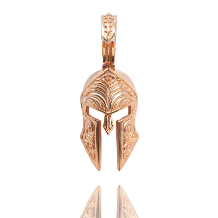 The Spartan Helmet Pendant - 14k Solid Gold is shaped like an ancient Greek warrior helmet with intricate engravings, shown upright on a white background with its reflection below.