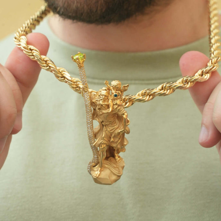 A person in a light green shirt holds a thick gold chain necklace featuring the Saint Christopher Pendant - 14k Gold, displaying an ornate robed figure with a jeweled staff topped with a green gem.