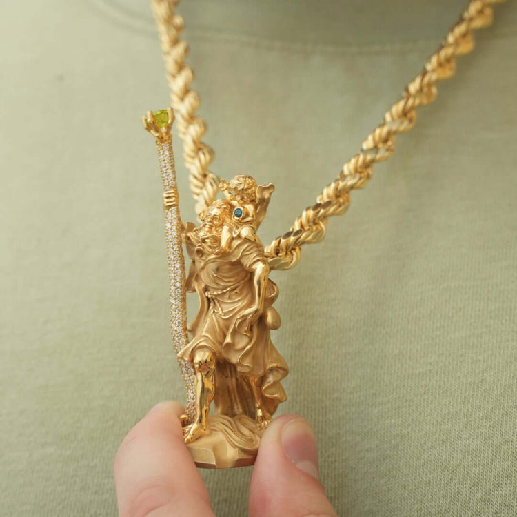 A person wearing a light green shirt holds the Saint Christopher Pendant - 14k Gold, featuring a detailed figure with a staff, hanging from a thick gold rope chain necklace.