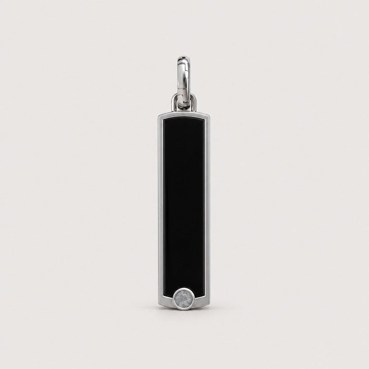 A rectangular black enamel pendant framed in 14k gold, adorned with a small round white gem at the base, hangs from a gold loop against a plain light background. This elegant piece embodies the timeless allure of the 14k Gold Black Enamel Tag Pendant with its sleek and sophisticated design.