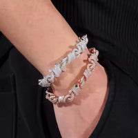 A person wears two Twisted Infinity Thorn Moissanite Bracelets—one in sterling silver and one in rose gold, each 15mm wide—on their wrist against a black fabric background.