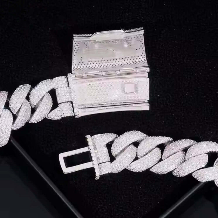 A close-up of the Moissanite Cross Cuban Link Chain - 18mm, a silver bracelet with a box clasp and sparkling VVS moissanite stones, displayed on a black textured surface.