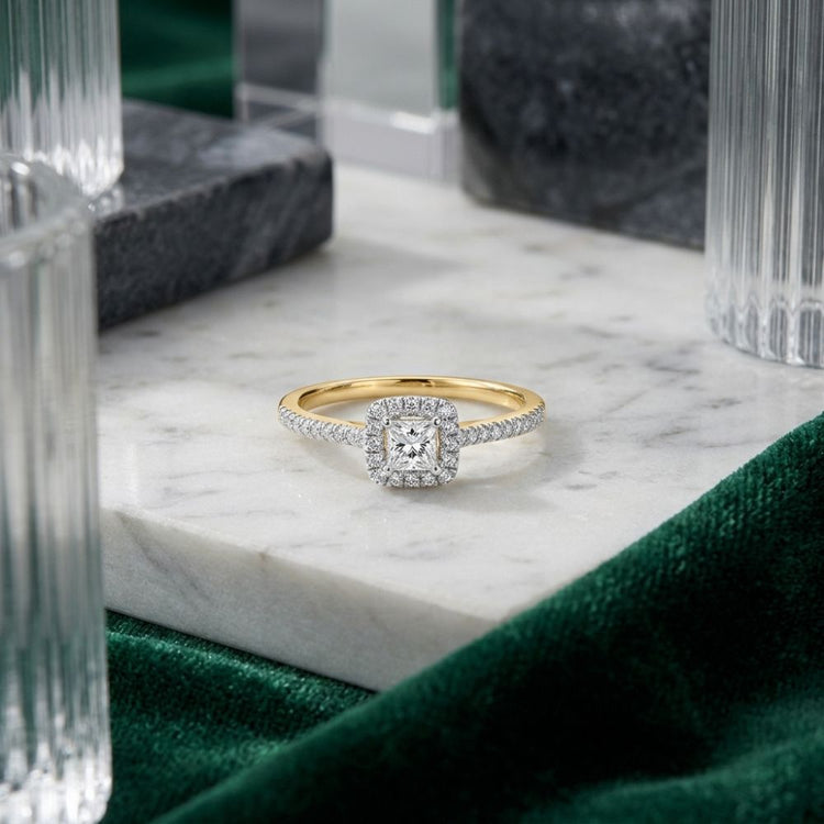 Showcased on a marble surface with glass and marble accents and green fabric, the 10k Gold Princess Diamond Halo Engagement Ring features a dazzling princess-cut diamond surrounded by a square halo of sparkling diamonds. This refined design adds timeless sophistication to any bridal engagement ring collection.