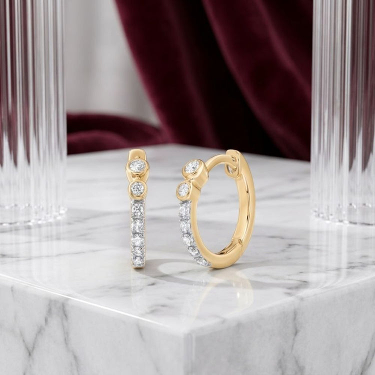 1/10 CTW Natural Diamond Hoop Earrings in 10K gold are displayed on a white marble surface, set against vertical glass columns and a burgundy velvet backdrop, highlighting the timeless elegance of these natural diamond hoop earrings.