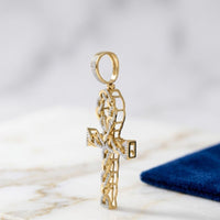 The 10K Gold Diamond Ankh Cross Pendant 3/8 CTW with diamond accents and braided details stands upright on white marble beside a dark blue textured fabric.