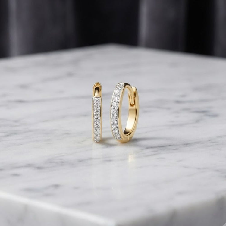 The 1/10ctw Natural Diamond Hoop Earrings in 10K Gold are shown encrusted with sparkling diamonds and elegantly displayed on a white marble surface, set against a dark, blurred background.