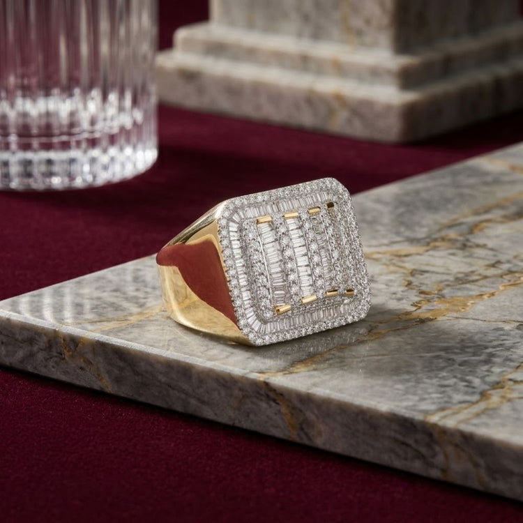 The 10k Yellow Gold Baguette Diamond Rectangle Ring, featuring a large rectangular face adorned with rows of sparkling diamonds, rests on a gray marble surface with a glass and stone object in the background. This eye-catching ring embodies elegance and modern style.