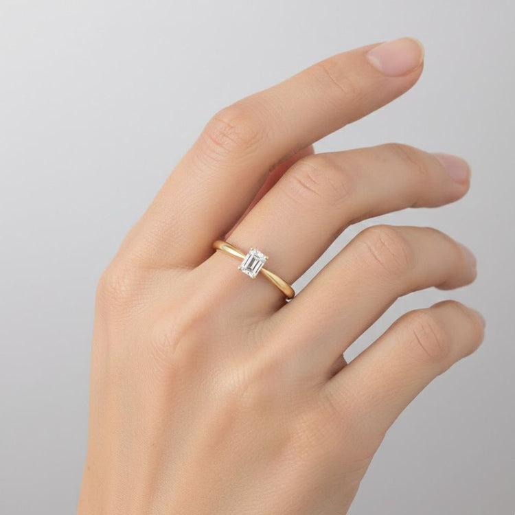 A hand adorned with the 1/4CT Diamond Emerald Solitaire Ring in 14K gold, set against a light gray background.
