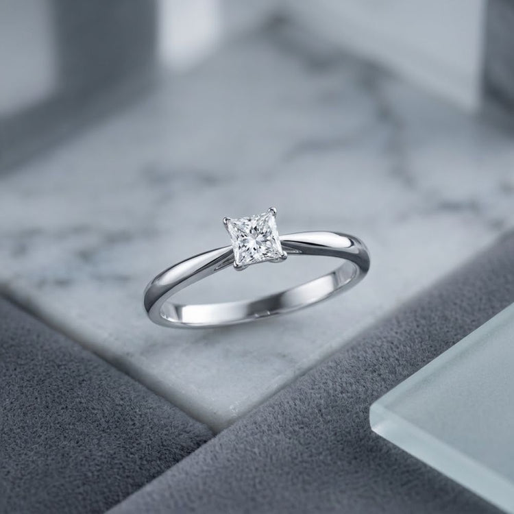 A beautiful 1/4CT Princess Diamond Solitaire Ring in 14K Gold with a square-cut natural diamond is displayed on a marble and gray geometric background.