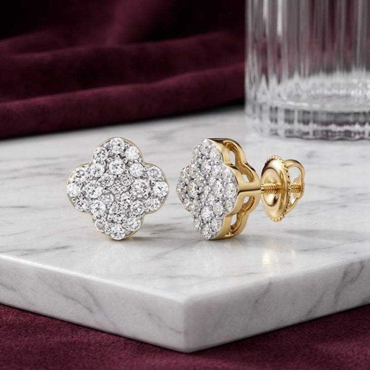 A pair of 1/6 CTW Natural Diamond Clover Earrings in 10K gold, adorned with sparkling natural diamonds, displayed on a white marble surface beside a faceted glass and burgundy fabric.
