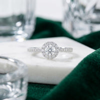 Experience the elegance of the 14k Gold Round Diamond Halo Engagement Ring, featuring a dazzling round center diamond encircled by a radiant halo of smaller diamonds. Set against a white marble backdrop and accented with touches of green fabric and glass, this exquisite engagement ring is the perfect symbol of your everlasting love.