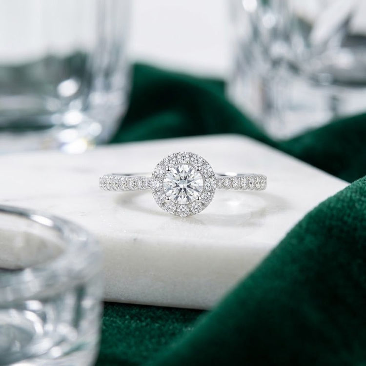 Experience the elegance of the 14k Gold Round Diamond Halo Engagement Ring, featuring a dazzling round center diamond encircled by a radiant halo of smaller diamonds. Set against a white marble backdrop and accented with touches of green fabric and glass, this exquisite engagement ring is the perfect symbol of your everlasting love.