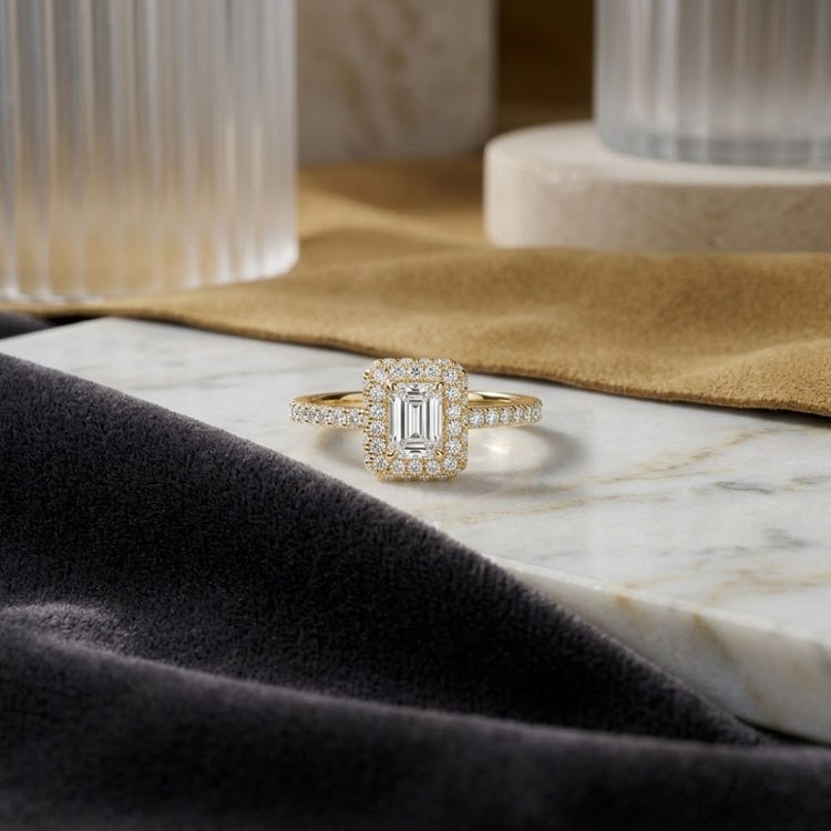 Displayed on a marble surface, the 14k Gold Emerald Diamond Halo Bridal Wedding Ring Set features a rectangular diamond encircled by a double halo of smaller diamonds. Surrounded by soft beige and dark gray velvet fabric, with glass and stone objects in the background, this stunning ring set captures timeless elegance—making it the perfect choice for any bridal wedding ring ensemble.