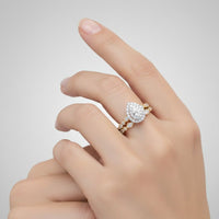 A hand with neatly manicured nails showcases the 10k Gold Pear Diamond Halo Bridal Ring Set 3/8 Cttw, featuring a pear-shaped diamond encircled by a halo of smaller diamonds on one ring and a coordinating band adorned with sparkling diamonds on the other, beautifully highlighting the set’s elegant design against a simple light background.