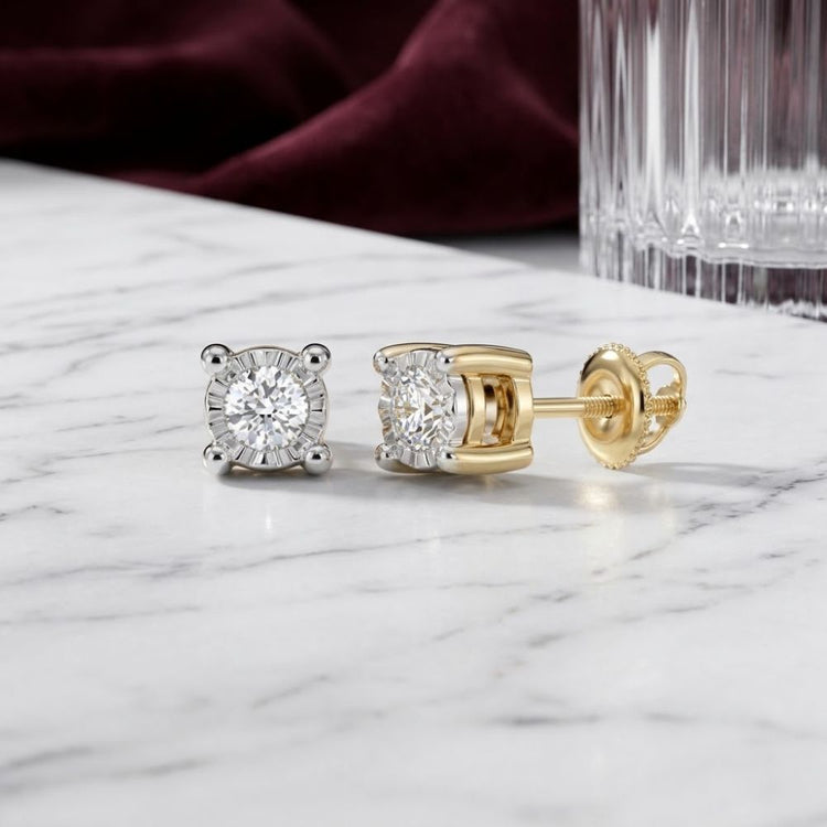 A pair of 10k Gold Round Diamond Stud Earrings 1/6 Cttw, featuring gold prongs and backs, is displayed on a white marble surface with a glass and burgundy fabric in the background.