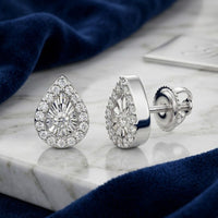 The 1/10CTW Natural Diamond Pear Earrings in 10K Gold feature a central stone surrounded by smaller diamonds, beautifully displayed on marble beside blue velvet fabric.