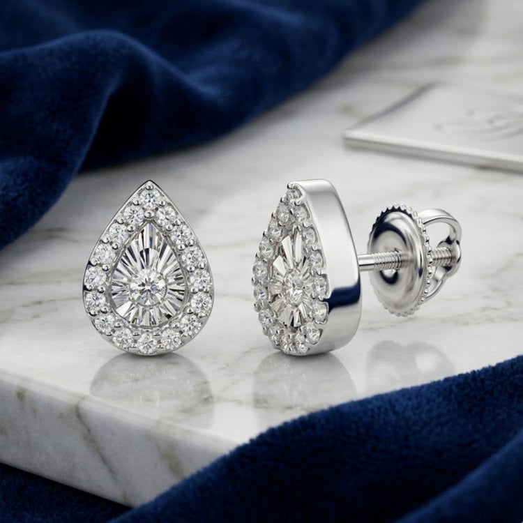 The 1/10CTW Natural Diamond Pear Earrings in 10K Gold feature a central stone surrounded by smaller diamonds, beautifully displayed on marble beside blue velvet fabric.