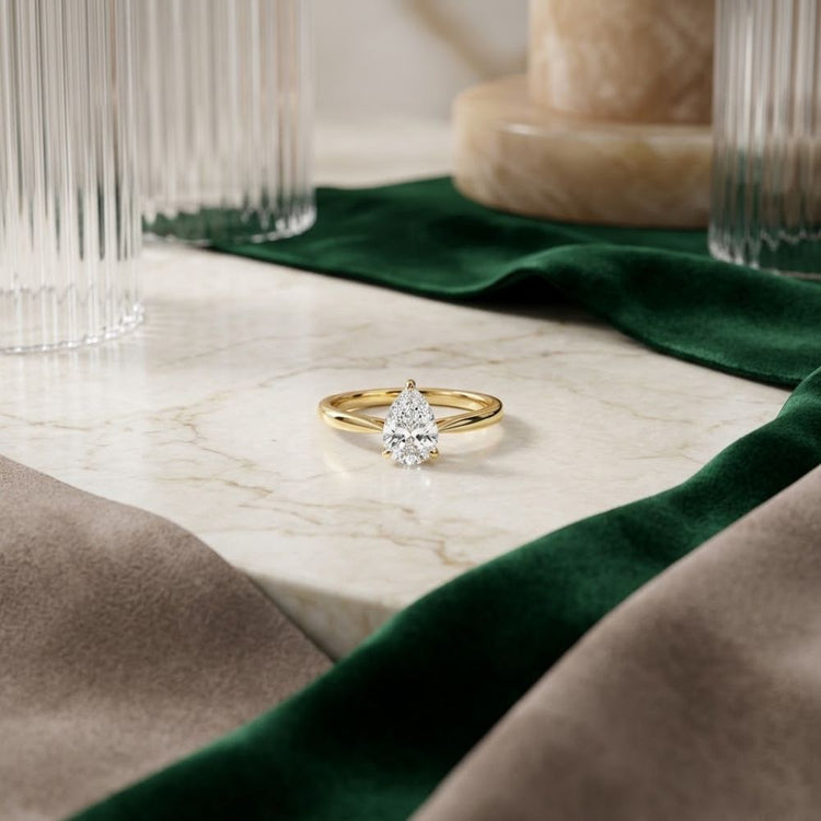 The 1/2CT Pear Diamond Solitaire Ring in 14k Gold is displayed on a marble surface, surrounded by green and taupe fabric, with decorative glass and marble objects in the background.