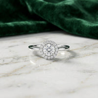 A 14k Gold Round Diamond Halo Engagement Ring rests elegantly on a white marble surface, its brilliance accentuated by green velvet fabric. This exquisite ring is an ideal choice for a timeless bridal engagement piece.