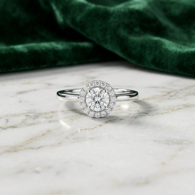 A 14k Gold Round Diamond Halo Engagement Ring rests elegantly on a white marble surface, its brilliance accentuated by green velvet fabric. This exquisite ring is an ideal choice for a timeless bridal engagement piece.