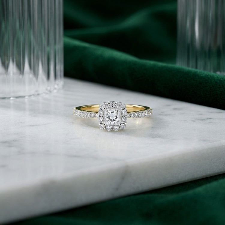 A 14k Gold Princess Diamond Halo Engagement Ring rests on a white marble surface, with green velvet and glass accents in the background.