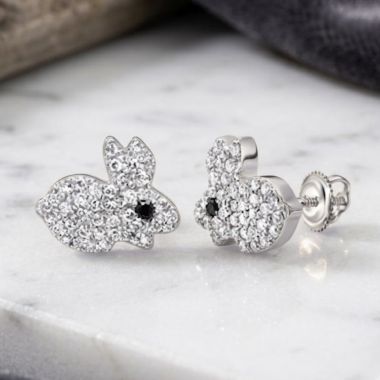 Two small, silver bunny-shaped earrings encrusted with clear gemstones and featuring striking black diamond eyes—these charming 10K Gold Black Diamond Earrings (1/4 CTW) are beautifully displayed on a marble surface.