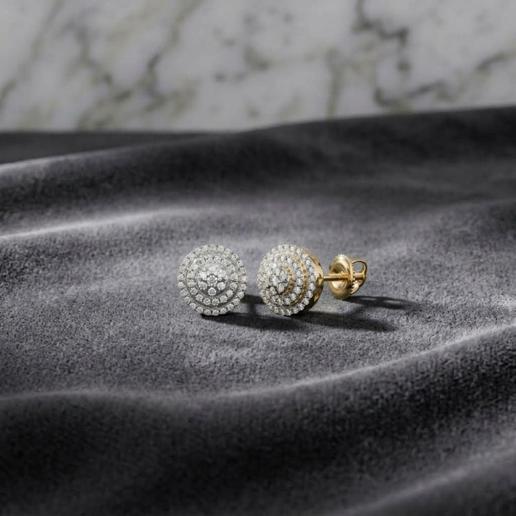 The 1/4 CTW Natural Diamond Round Earrings in 10K gold are elegantly showcased on a soft gray velvet surface, with their sparkling natural diamond studs set against a blurred marble background.