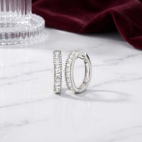A pair of 10K Gold Round Diamond Hoop Earrings 1/4 CTW, adorned with small sparkling stones, rests on a white marble surface, complemented by ribbed glass and folded burgundy fabric in the background.