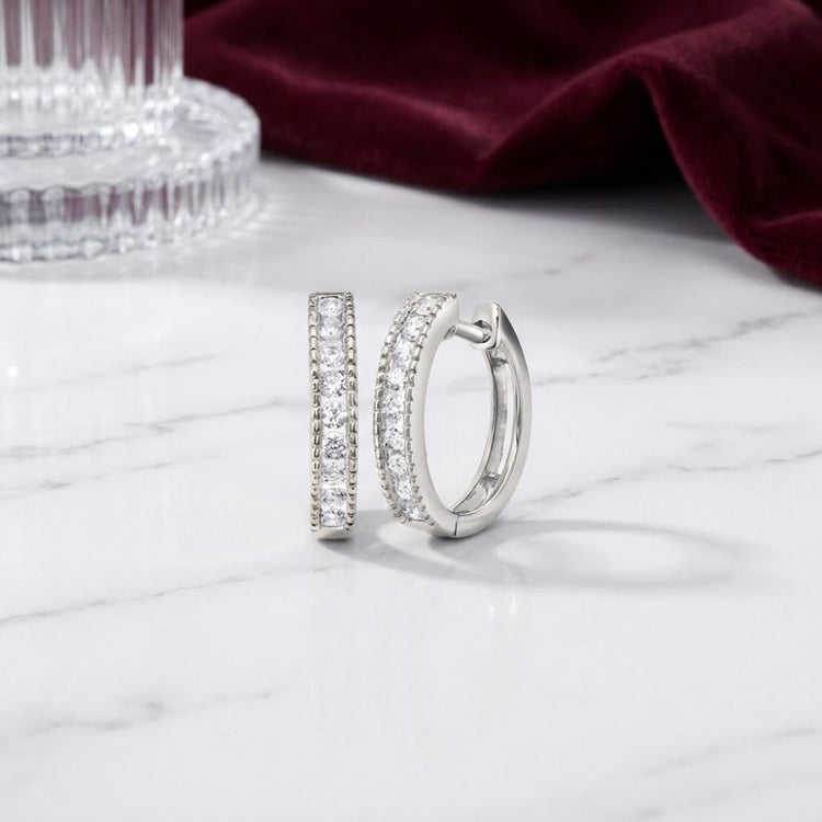 A pair of 10K Gold Round Diamond Hoop Earrings 1/4 CTW, adorned with small sparkling stones, rests on a white marble surface, complemented by ribbed glass and folded burgundy fabric in the background.