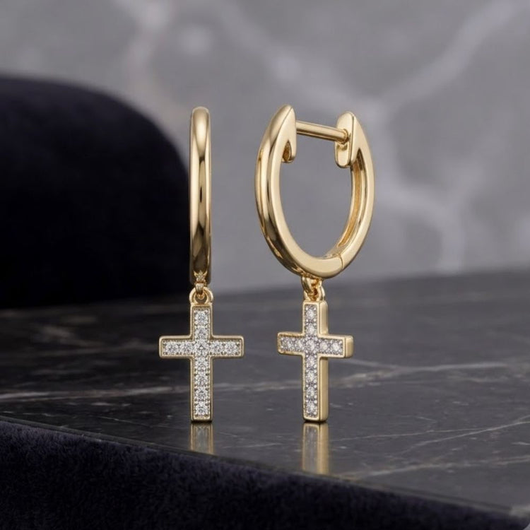 Two 1/20CTW round diamond cross dangle hoop earrings in 10K yellow gold from Nicole’s Dream Collection displayed on a dark marble surface with a soft, blurred gray background.