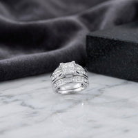 A breathtaking 14K Gold Princess Diamond Bridal Ring Set (2 CTTW) features a square-cut center stone accented by sparkling diamonds, presented on a marble surface with a dark fabric and stone block in the background.