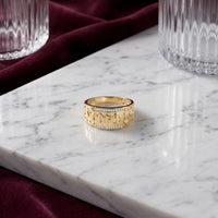 A 10k Gold Dad Men’s Ring with Natural Diamonds featuring intricate patterns and a central row of sparkling stones rests on a white marble tray, surrounded by textured glass cups and a burgundy fabric.