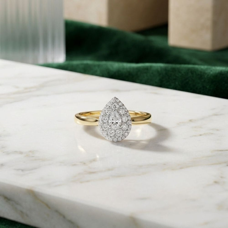A 14k Gold Pear Diamond Halo Engagement Ring rests on a white marble surface, with green and beige background elements.