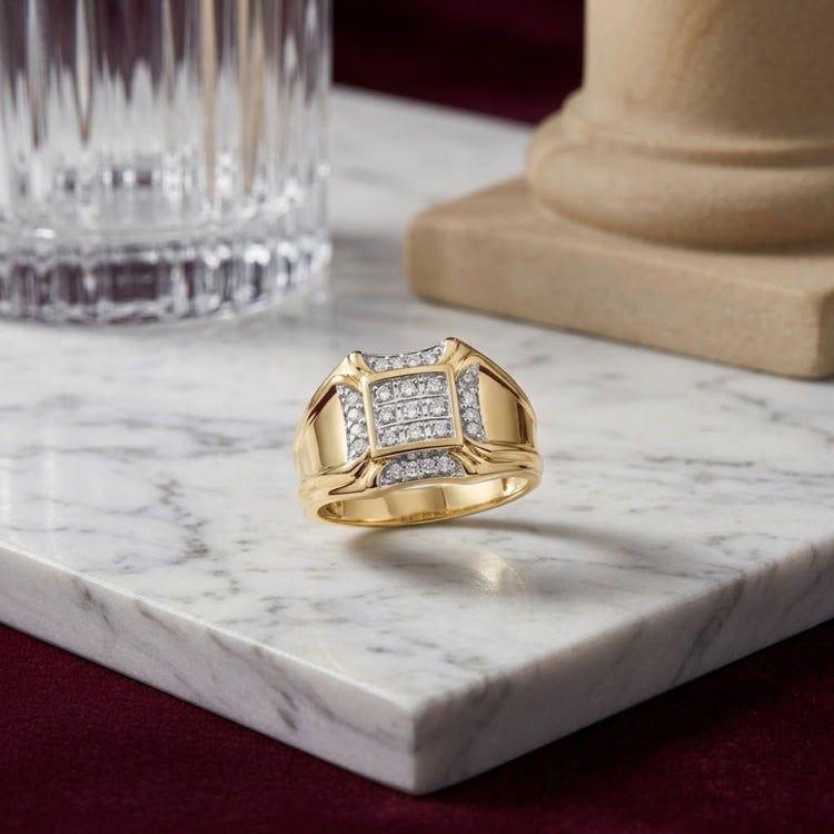 The 10K Yellow Gold Diamond Cluster Ring 1/4CT, featuring square and rectangular diamond accents, is displayed on a marble surface beside a glass and the base of a stone column, set against a burgundy fabric background.
